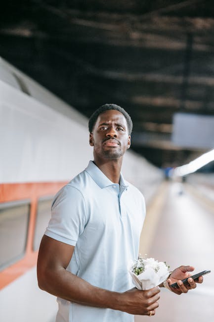 A young man with dark skin, short black hair, and a serious expression, stands on a platform or station with an arched ceiling, holding a small bouquet of white roses wrapped in white paper in one han